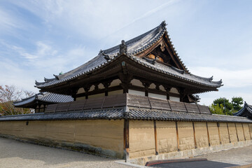 Fototapeta premium Wide view of Gomado Hall at Horyuji temple in Nara, featuring layered earthen walls and an elaborate wooden roof with decorative tiles under a bright sky. The historic architecture is highlighted by