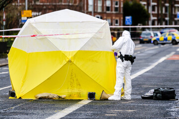 A scene of crime officer wearing a white boiler suit looks inside an examination tent at a crime scene © Stephen