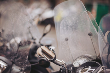 Scooters park closely together, their clear windshields reflecting droplets of recent rain. The atmosphere is alive with the sounds of the city on a cloudy afternoon