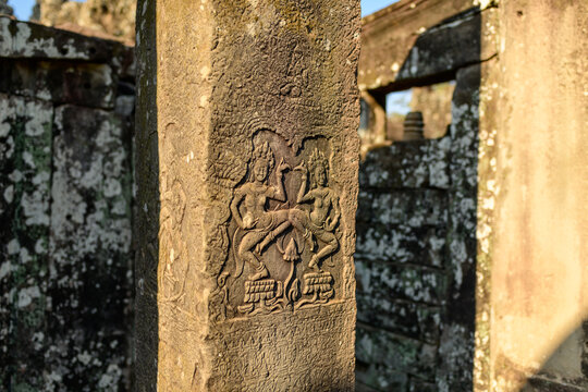 Detailed stone relief of two apsara dancers adorns a weathered sandstone pillar at Bayon temple in Angkor, Cambodia. Warm sunlight brings out the intricate Khmer carvings and textured surfaces, with
