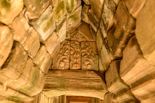 A close-up of a weathered sandstone lintel reveals detailed floral carvings within the ancient corridors of Baphuon temple in Angkor, Cambodia. Warm light highlights the textured stone and intricate