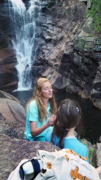 Happy mother and daughter talking by a forest waterfall in Norway. Bright northern daylight, authentic family bonding and sustainable travel concept.