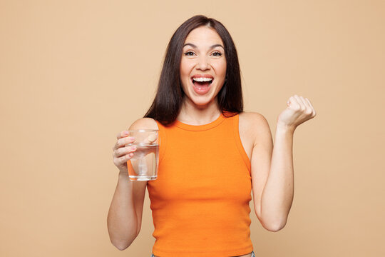 Young woman wear orange casual clothes hold in hand glass drink water do winner gesture isolated on plain pastel light beige background. Proper nutrition healthy fast food unhealthy choice concept...