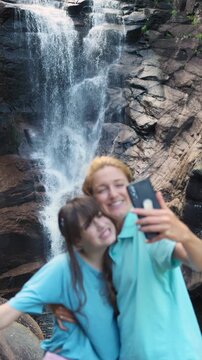 Happy mother and daughter taking a selfie by a forest waterfall in Norway. Authentic family connection, joy, and nature wellness during a summer hike in Northern Europe.