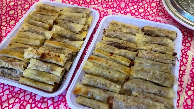 Close-up of freshly prepared crispy meat spring rolls neatly arranged in plastic trays on a red patterned tablecloth, ready to serve at a gathering or family meal.