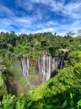 A breathtaking waterfall cascades down a lush green cliff into a deep valley, surrounded by tropical forest under a bright blue sky with scattered clouds, creating a serene and dramatic natural panora