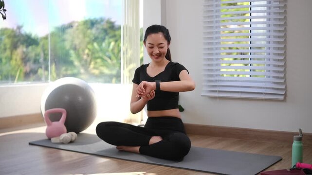 Smiling woman checking smartwatch while sitting on yoga mat at home. Fitness training, and healthy lifestyle concept.