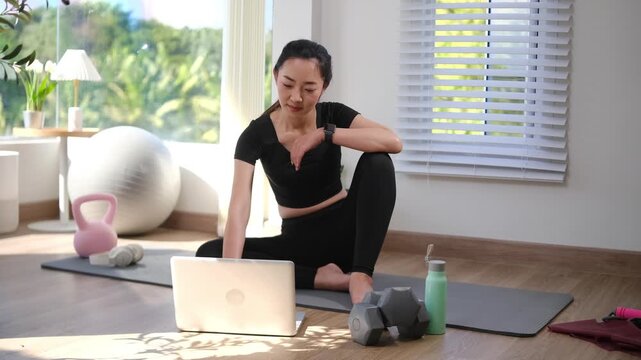 Smiling woman sitting on yoga mat using laptop after home workout. Home fitness and healthy lifestyle concept.