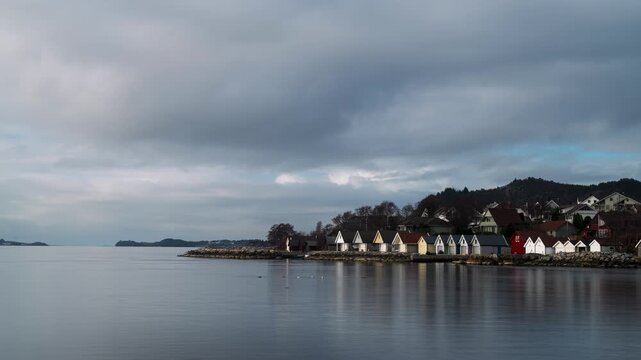 Vibrant a-framed boat houses next to the ocean with mountain tops in the background, in a small Norwegian costal town. Clouds moving on the sky, birds swimming on the water in the foreground.