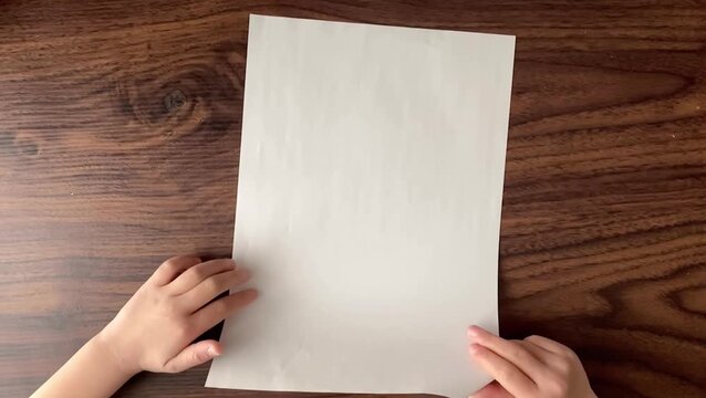 Child folding A4 paper on wooden desk, top view, slow motion, no sound