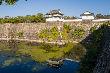 Traditional white castle turrets rise above ancient stone walls, their shapes mirrored in the tranquil moat below. Lush greenery and bright sunlight create a vivid, peaceful scene in Osaka, Japan. © Florent