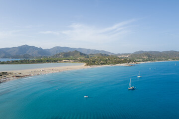 Fototapeta premium Crystal-clear turquoise sea with anchored sailboats stretches toward a wide sandy beach and lush hills, with rugged mountains in the distance near Porto Giunco.