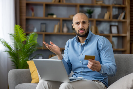 Young man sitting on a sofa, looking confused and shrugging while holding a credit card and using a laptop, experiencing difficulties with online banking or shopping