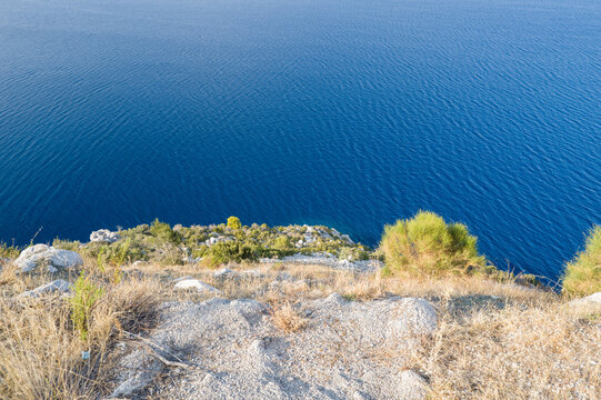 Sunlit rocky bluff with dry grasses and sparse green shrubs overlooks the calm, intensely blue sea along the Argolide coast in Peloponnese.