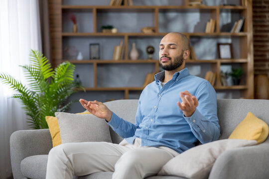 Young man practicing mindfulness meditation on a comfortable sofa in his living room, focusing on breath and inner peace for relaxation, stress relief, and improved mental health at home