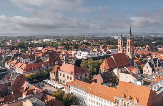 Aerial drone view of Goettingen (G&ouml;ttingen) old town in Germany, featuring red-roofed historic buildings and church towers, with a small statue visible near a public square under a partly cloudy sky.