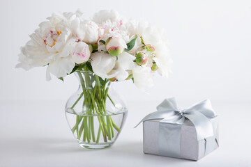 Obraz premium White peony flowers arranged in a clear glass vase next to an elegant gift box with ribbon on a white background. Romantic floral still life for celebration, wedding, and luxury gift concepts.