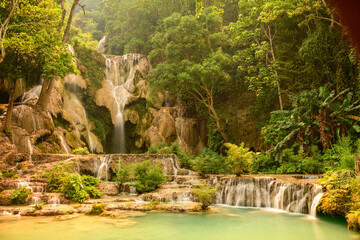 Warm sunlight bathes the layered limestone cascades and tranquil turquoise pools of Kuang Si Waterfalls, surrounded by dense tropical forest in northern Laos. Lush greenery and flowing water create a © Florent
