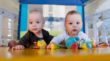 Two adorable twins are on their bellies in a lively playroom, surrounded by an array of colorful toys. Their curious expressions show excitement as they interact with their playful environment