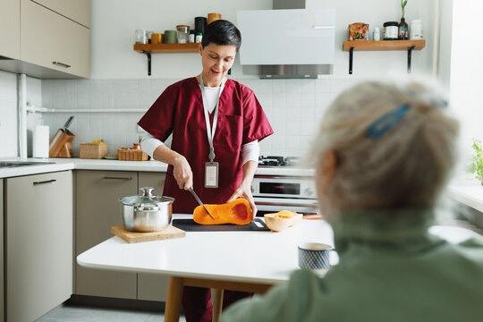 Selective focus on nurse social service volunteer cutting pumpkin at kitchen table to prepare healthy breakfast for old lady, while talking to her patient sitting with back turned to camera