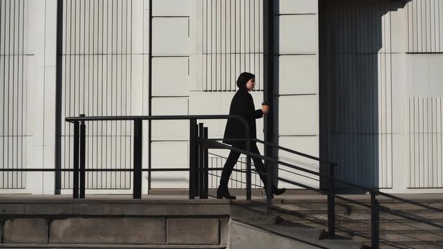 European woman walking along a modern building holding a coffee cup, transitioning from a shadowed area to a well-lit section of the urban environment