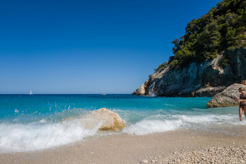 Obraz premium Bright turquoise waves splash against sunlit rocks on a pebbled beach, with dramatic limestone cliffs and lush greenery framing the cove at Cala Goloritze, Sardinia. Clear blue sky and vivid water