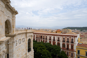 Fototapeta premium Detailed stonework of the Bastione di Saint Remy rises above leafy trees and historic buildings, with Cagliari's cityscape and distant hills under a softly clouded sky.
