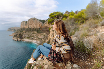 Female hiker with backpack and hat stands on top of cliff, admiring seascape. A young woman with a backpack feels free, enjoying the view. View from behind. Concept of hiking, freedom, and nature.