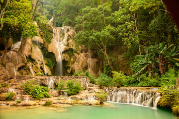 Warm golden light illuminates the multi-level limestone cascades and turquoise pools of Kuang Si Waterfalls in northern Laos. Lush tropical forest surrounds the flowing water, creating a vibrant and © Florent