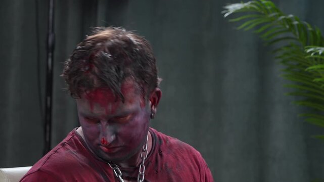 Close-up of a man with blue and purple zombie makeup on his face, looking up with a tense dramatic expression, wearing a red shirt and a massive chain around his neck, against a dark studio background