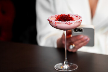 A close-up of an elegant pink cocktail in a coupe glass with a sugared rim and rose garnish, held...