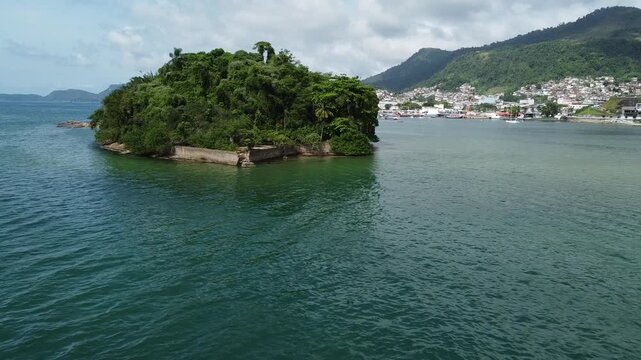 Drone footage from low altitude with the drone approaching the Island Ilha do Coqueiro in Angra dos Reis. The center of town the forest covered mountains, as well as the harbour is in the background