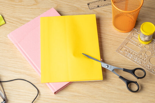 A yellow sheet of paper is being cut with scissors on a wooden desk alongside a pink notebook and school supplies.