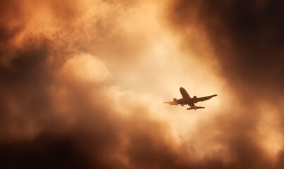 An airplane soars through dramatic, golden-orange clouds during sunset or sunrise, creating a striking silhouette against the vibrant sky.