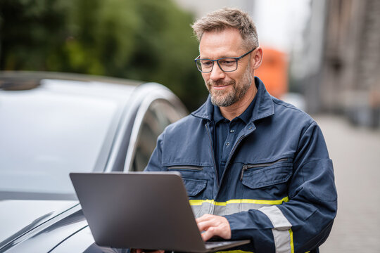 A man in a work uniform uses a laptop while standing next to a car outdoors, focused on his task.