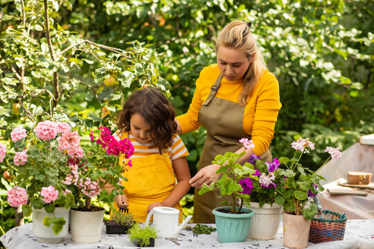 A woman mom and a kid girl child carefully tend to vibrant potted flowers in a lush garden, wearing gardening gloves and aprons, surrounded by greenery and blooming plants.