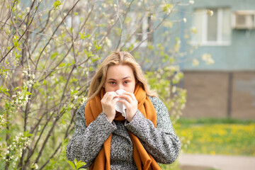 A young woman with a mustard scarf and cozy sweater blows her nose outdoors amidst blooming spring branches, suggesting seasonal allergies or a cold.