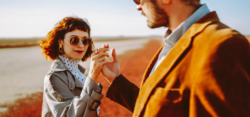A couple stands together in a field overgrown with bright orange and yellow plants. They smile and embrace, enjoying the warm autumn sun. The photo features a clear sky and an open landscape. © maxbelchenko