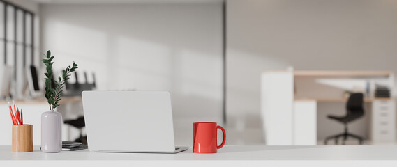 Modern 3D office desk with laptop, red mug and plant in blurred open workspace