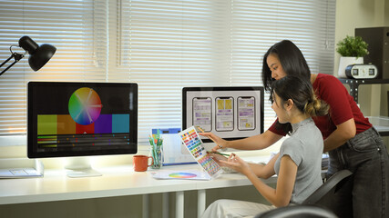 Two designers collaborate on mobile app UX UI, reviewing color palettes and wireframes on desktop screens in a modern creative office