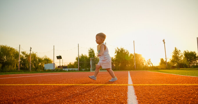A young boy runs around a tennis court, happily kicking a ball. The sun sets in the background, creating a warm atmosphere as he enjoys his playtime.