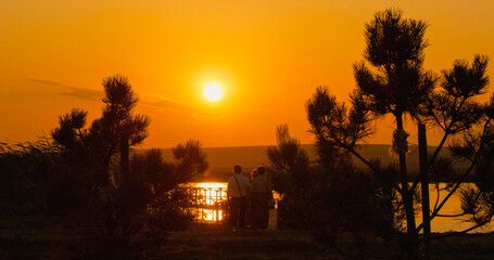 People stroll along a picturesque lakeside toward a pontoon as the sun sets, casting a warm glow over the serene landscape and reflecting on the water's surface. © RecCameraStock