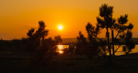People stroll toward a pontoon near the lakeside, framed by trees, as a stunning sunset casts warm colors across the sky, creating a serene atmosphere. © RecCameraStock