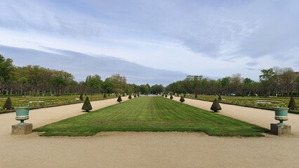 There are many flower beds in the park next to dirt paths and grassy lawns. Tulips, daffodils and other flowers grow on them. Various trees grow nearby. Sunny spring weather and cloudy sky