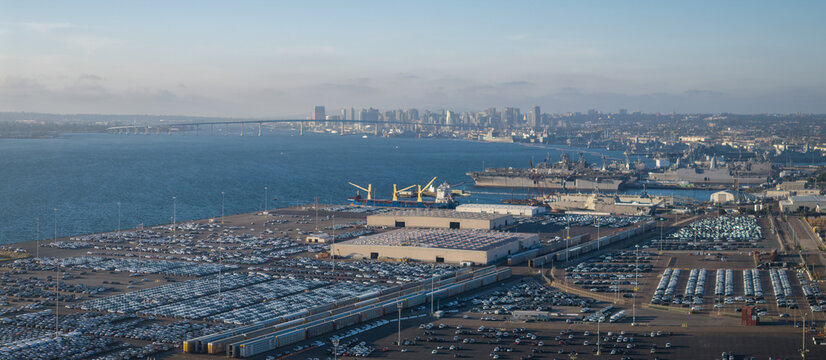 Aerial view of the Coronado bridge stretching over the bay towards the city skyline, with ships docked and cars parked in organized rows, San Diego, California, United States.