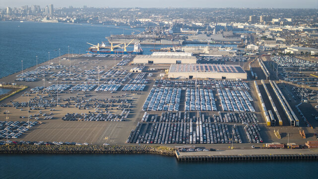 Aerial view of vast rows of cars create a geometric tapestry on the port, contrasting with the blue waters of the bay and industrial structures in the distance, San Diego, California, United States.