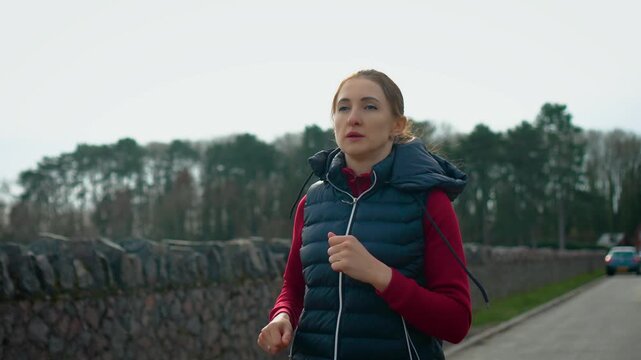 Wellness focused woman running near stone wall by lakeside during morning session. Determined female athlete exercising outdoors enhancing endurance and physical fitness. Healthy living concept