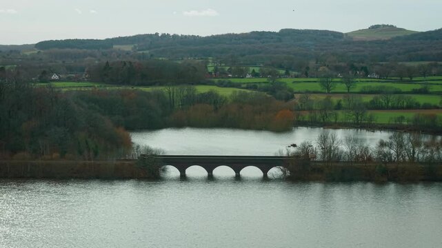 Stone arch bridge stretching across calm lake surrounded by leafless trees. Rolling hills rising beyond green fields and scattered rural houses. Overcast sky hanging above tranquil countryside with