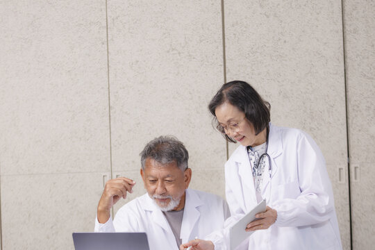 Elderly male and female doctors in white coats discussing medical information on laptop and tablet in clinical setting. They appear focused and engaged in their work