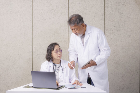 Two doctors in white coats are discussing medical information at desk. One is seated with laptop, while other stands holding tablet. They appear focused and engaged in their work